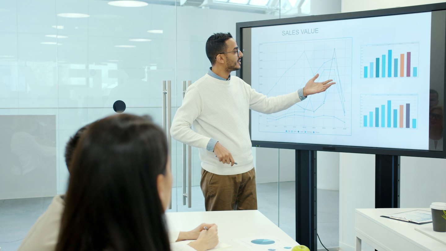Man presenting sales data on screen to colleagues in a meeting room.